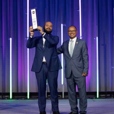 two men in suits standing on stage together, one holding a trophy up at shoulder length