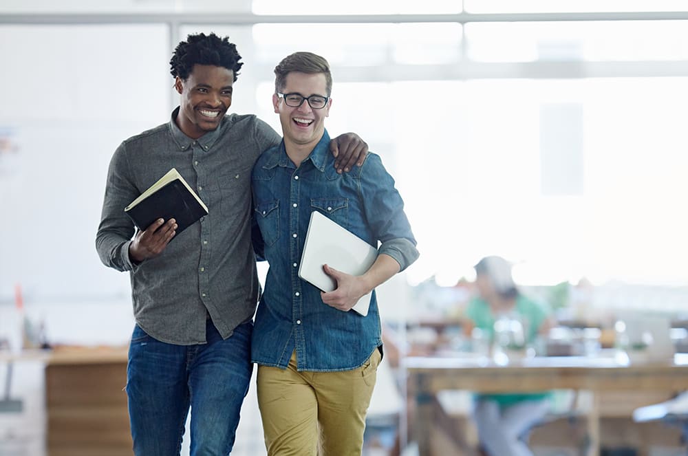 A man holding a notebook laughing with a man holding a laptop