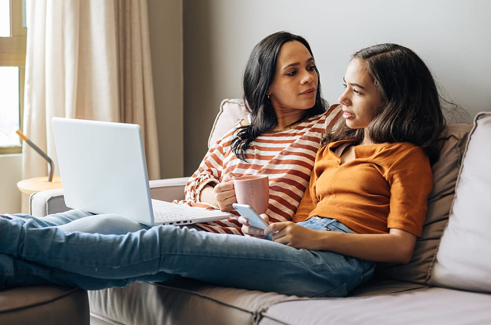A woman holding a cup sitting on a couch next to a young girl holding a phone