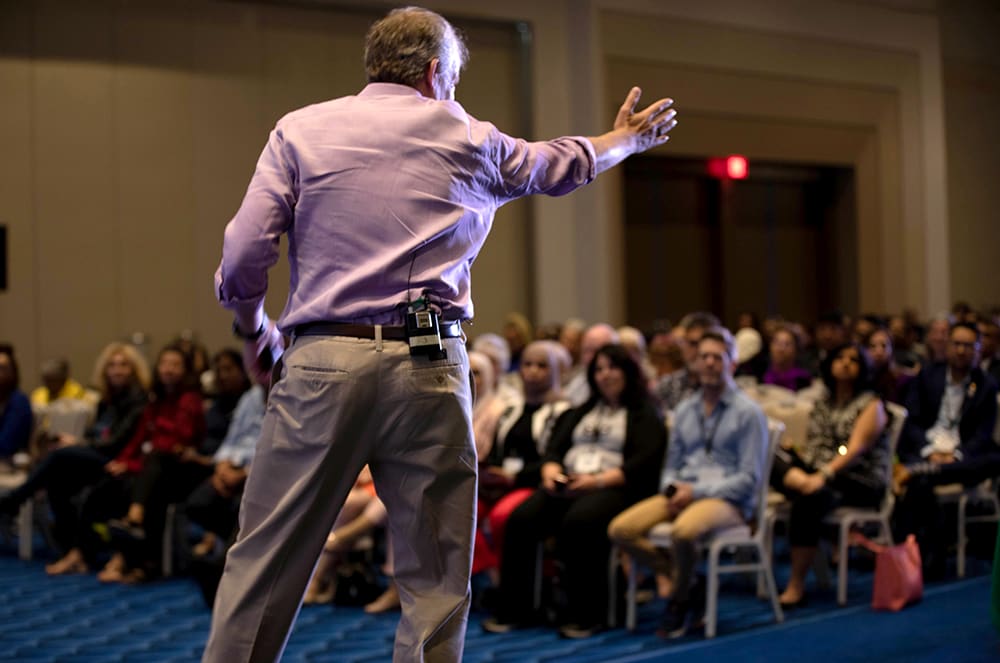 Man in pink shirt with arm outstretched speaking to an audience