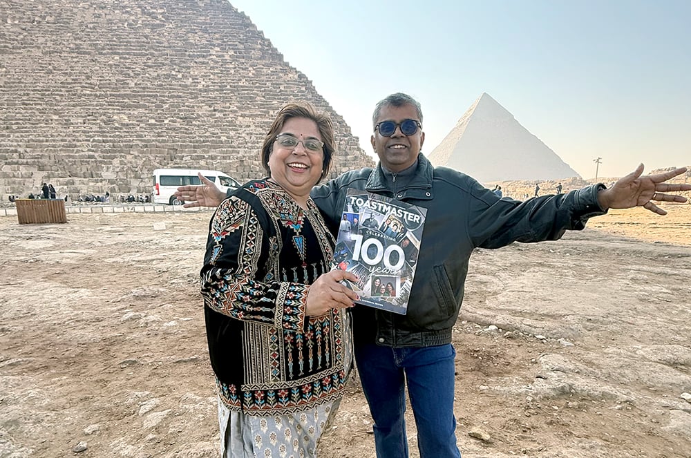 Woman holding a magazine and a man with his arms outstretched standing in front of the Pyramids of Giza