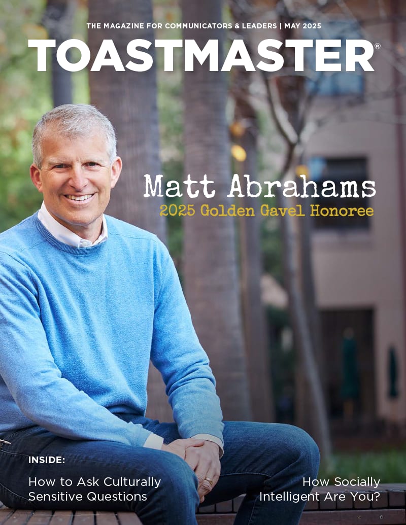 The image features a smiling older man with gray hair wearing a blue sweater, sitting outdoors in front of a building with trees in the background.