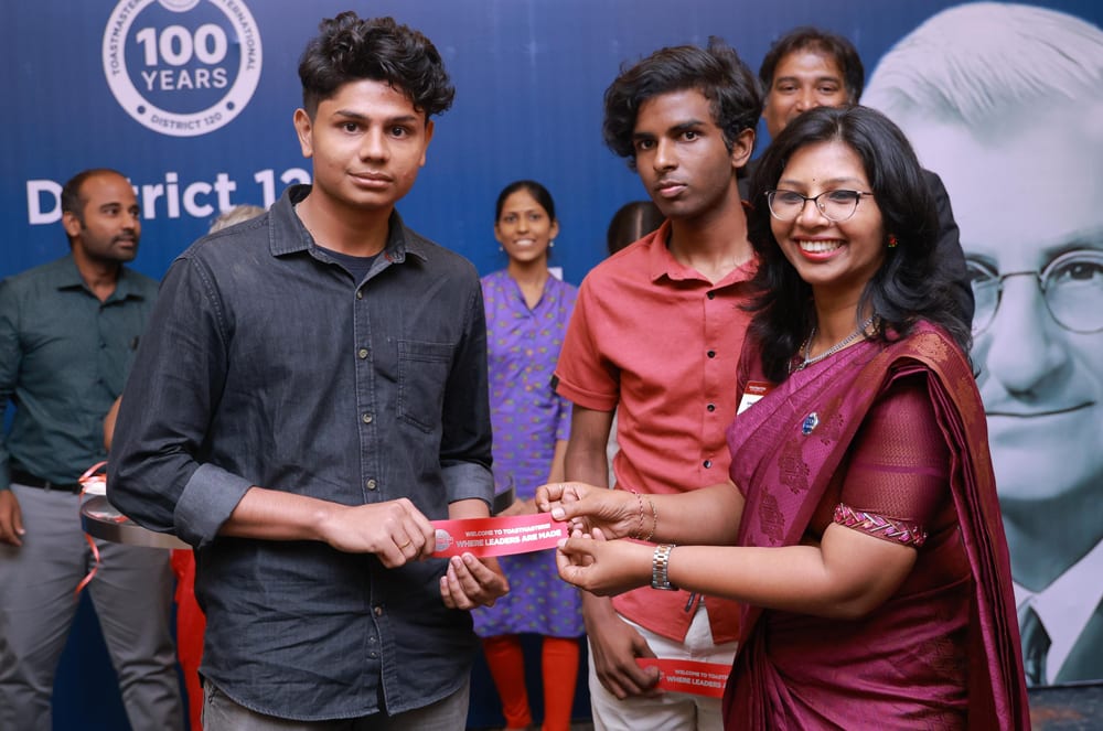 A man and woman holding a red Toastmasters ribbon with a group of people smiling behind them