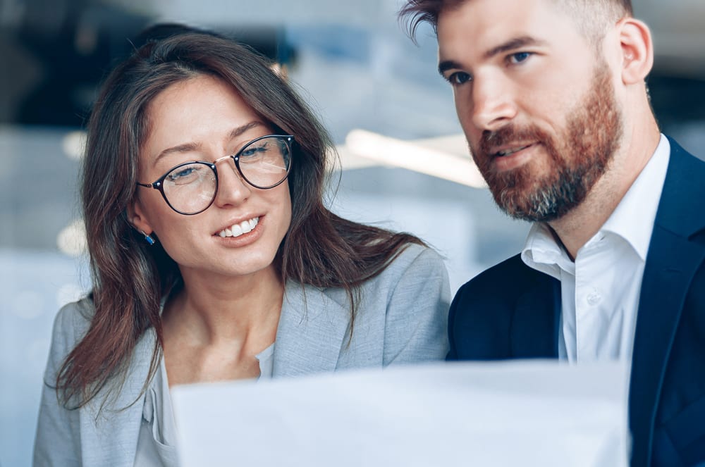 The image shows a smiling young woman wearing glasses and a man with a beard, both dressed in business attire, standing together in what appears to be a professional setting.