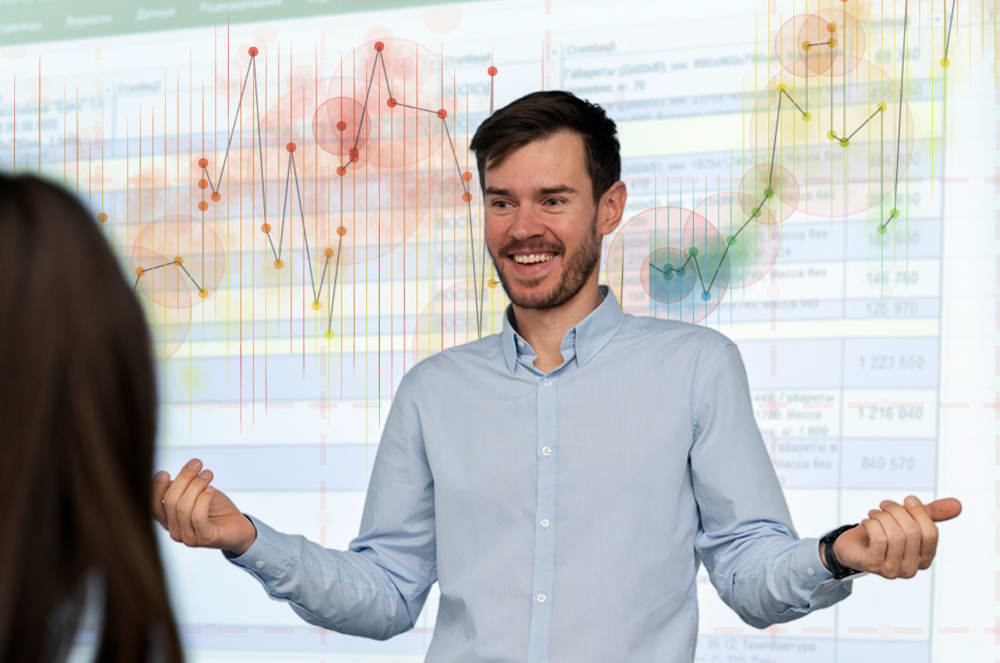 A smiling man in a light blue shirt stands in front of a whiteboard with various graphs and charts displayed on it.