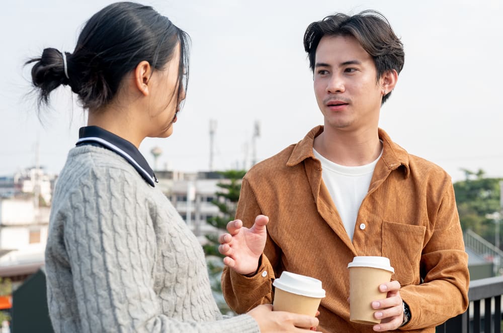 Two young people, a man and a woman, are engaged in conversation while holding coffee cups outdoors against a blurred urban background.