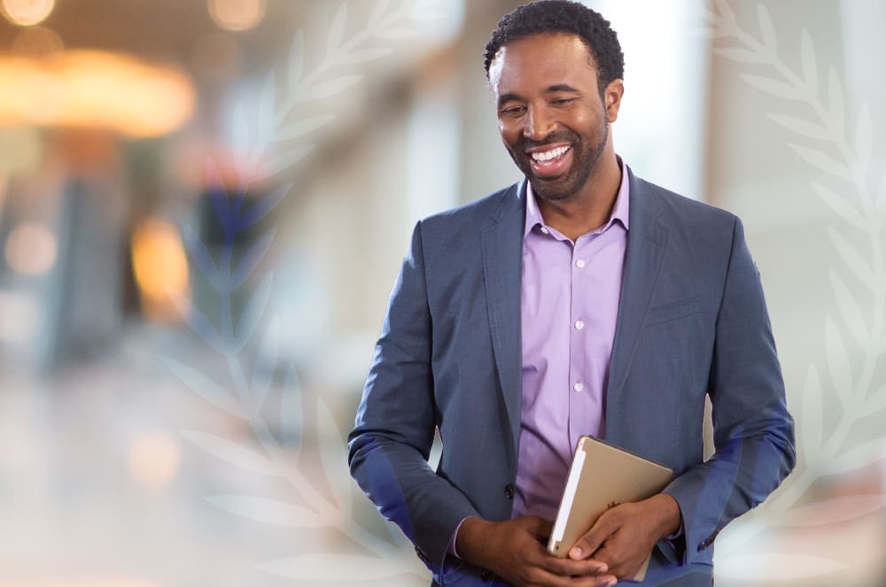 A smiling man in a gray suit and pink shirt stands in a blurred urban setting, holding a laptop or tablet in his hands.