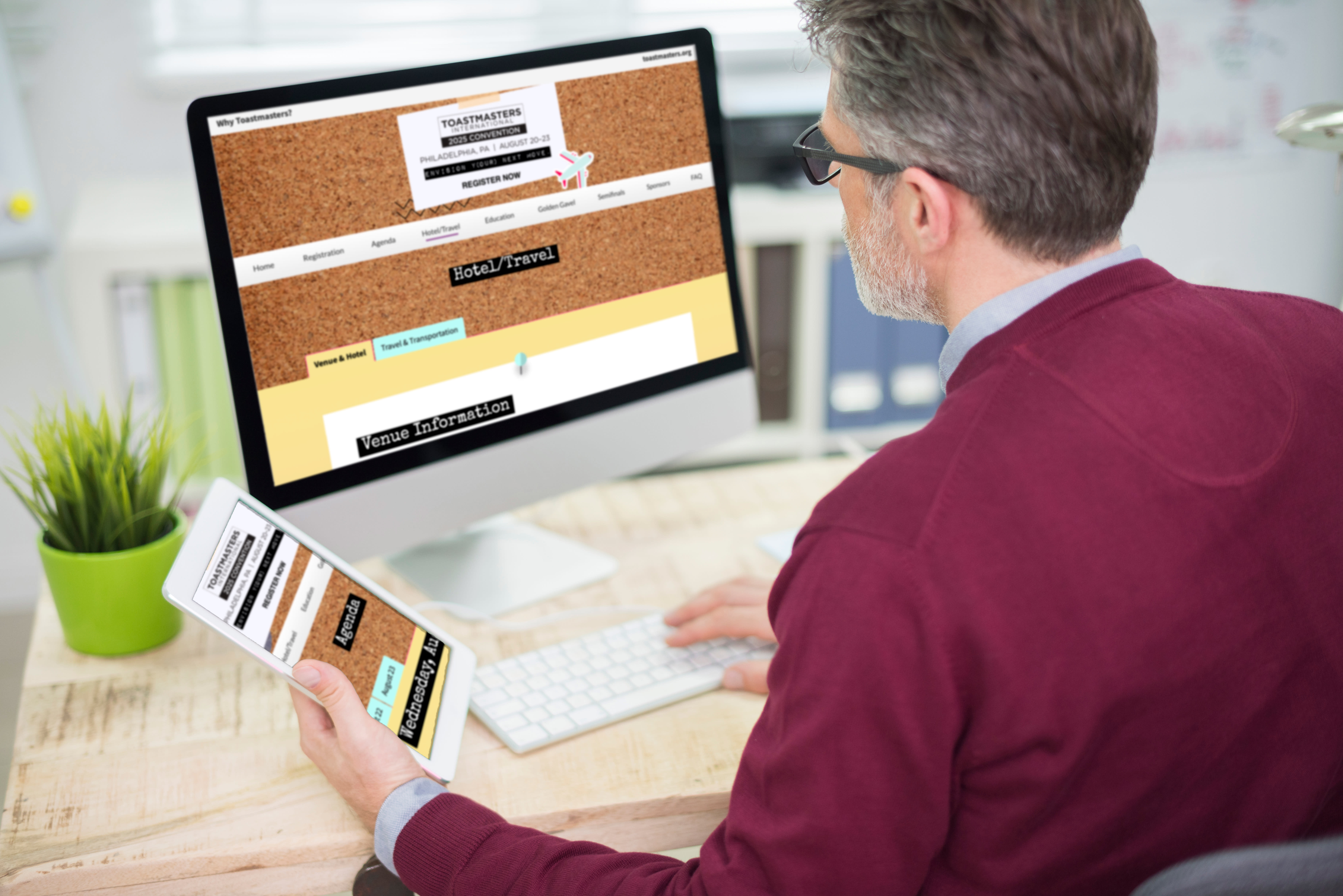 A man in a burgundy shirt is sitting at a desk, working on a computer with a corkboard-style website displayed on the screen.