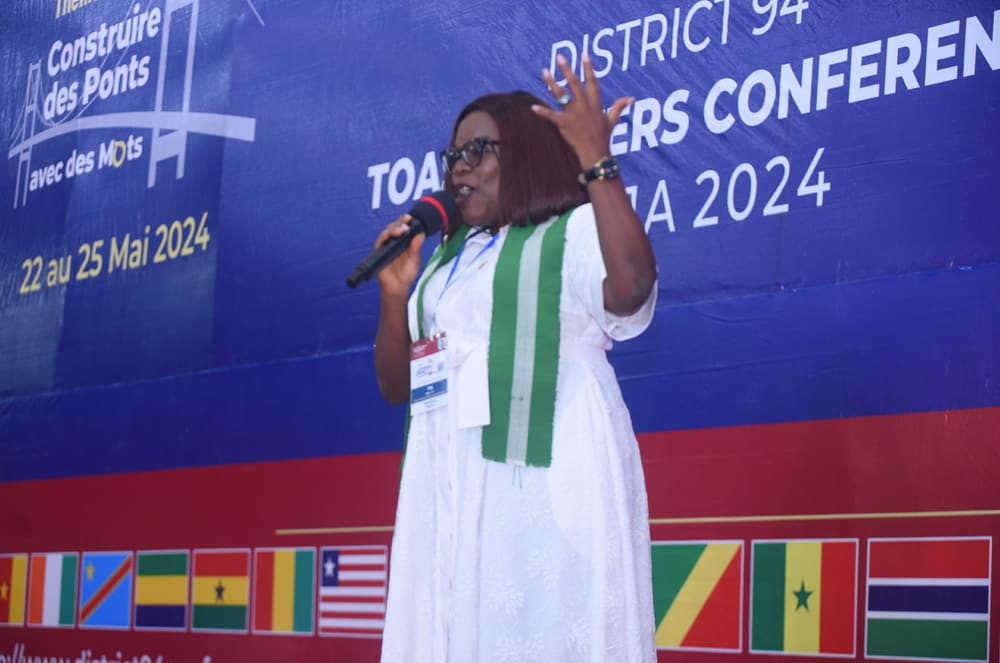A woman in a white dress with green and white stripes is speaking into a microphone on a stage, with a backdrop featuring the flags of various countries.