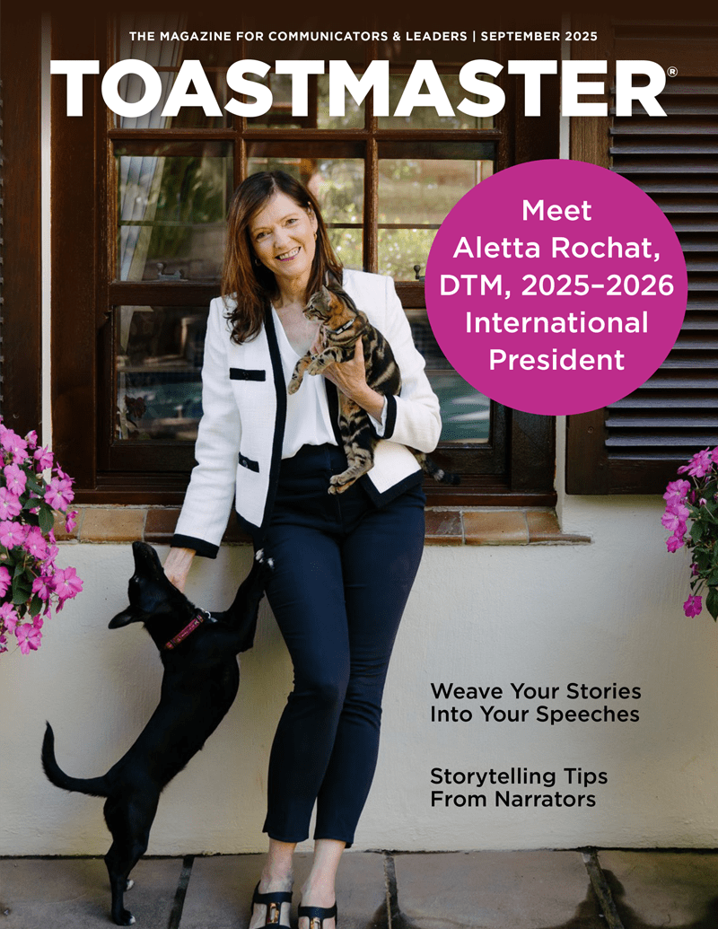 The image shows a smiling woman in a white jacket and jeans standing in front of a building with pink flowers in the background.