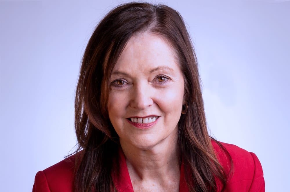 A smiling woman with long brown hair wearing a red top against a light blue background.