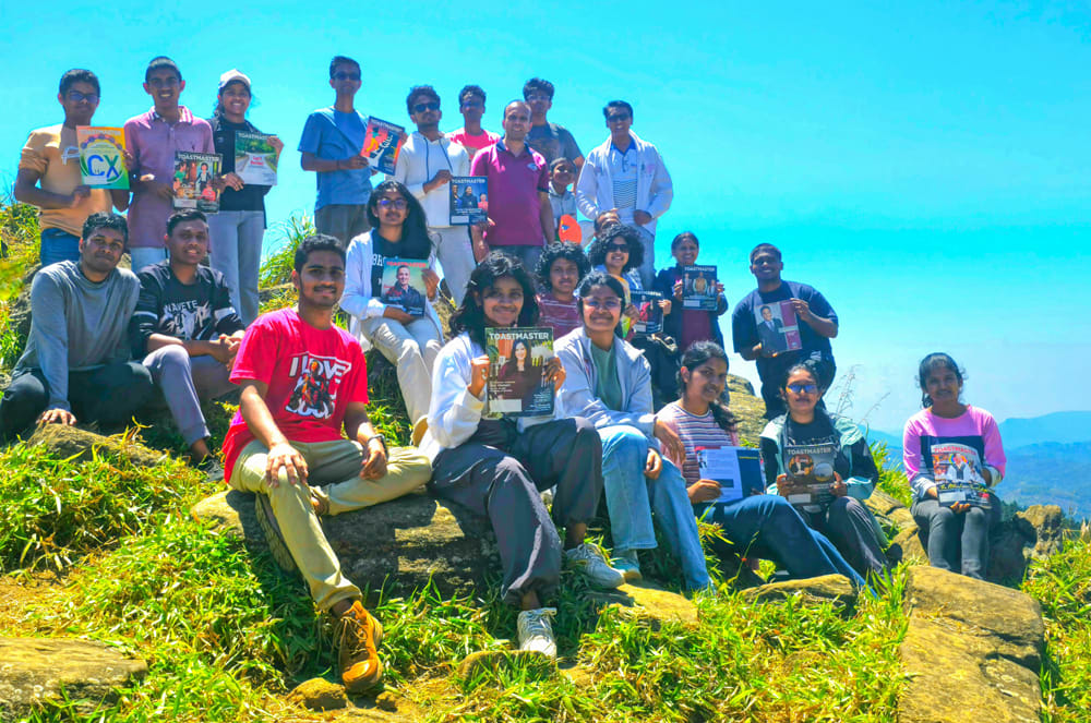 A group of people, both adults and children, sitting together on a grassy hill with a bright blue sky and mountains in the background. : Group of people holding magazines posing on a grassy mountain