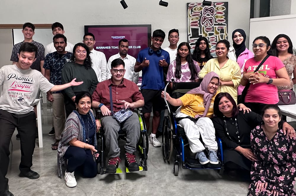A group of diverse individuals, some seated and some standing, gathered in what appears to be a community or office setting, with various decorations and signage visible in the background. Mawaheb Toastmasters Club, art studio, Toastmasters club, Toastmasters members, club designed for intellectual disabilities