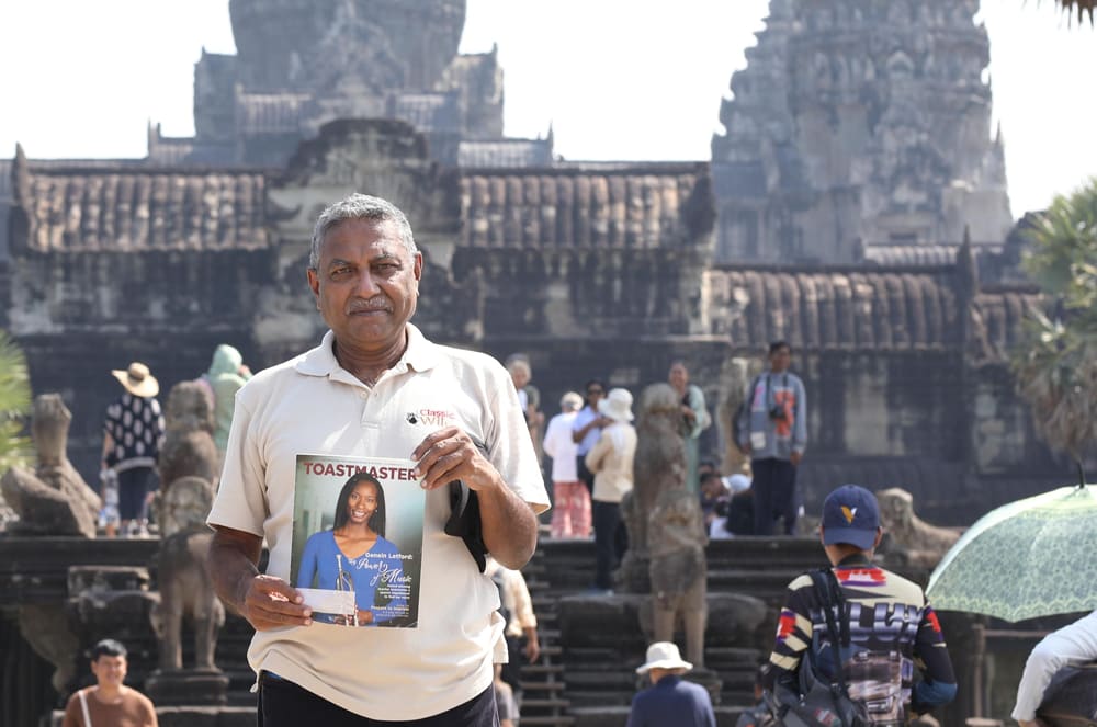 An elderly man in a white shirt stands in the foreground, holding a Toastmasters book, with a large, ornate temple structure visible in the background.