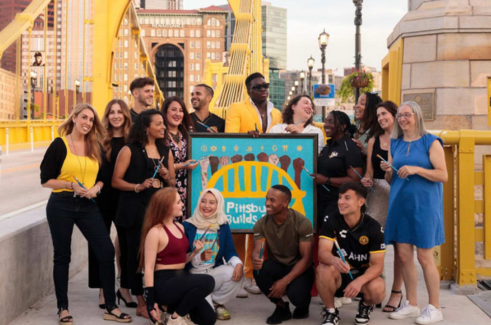A group of diverse individuals, including men and women of various ages and ethnicities, are gathered together in an urban setting with colorful buildings and structures in the background. Photo credit: Dominique Murray