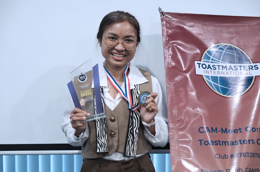 A smiling woman in a white coat and striped shirt stands in front of a Toastmasters International banner, holding several awards and medals.