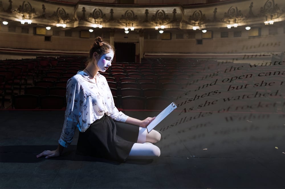 A person is kneeling on the stage of a dimly lit theater, with text projected on the wall behind them.