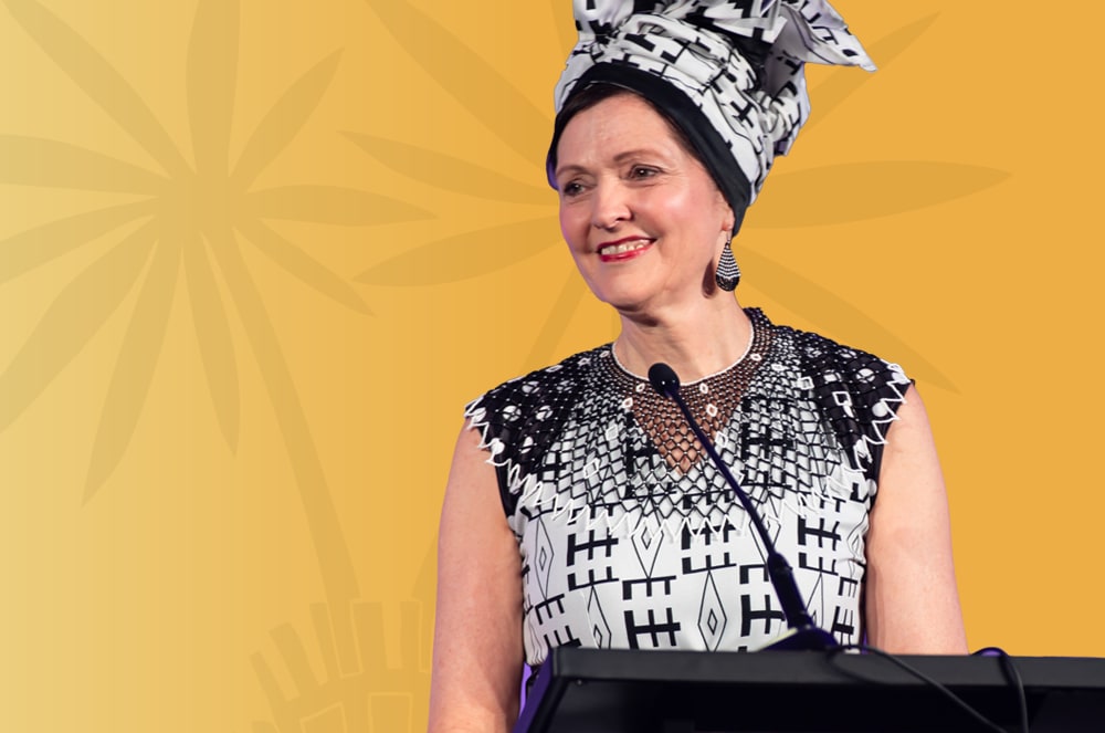 A smiling woman wearing a patterned headscarf and dress stands in front of a bright yellow background. Aletta Rochat chaired the 2024 World Championship of Public Speaking. She wore South African-themed attire.