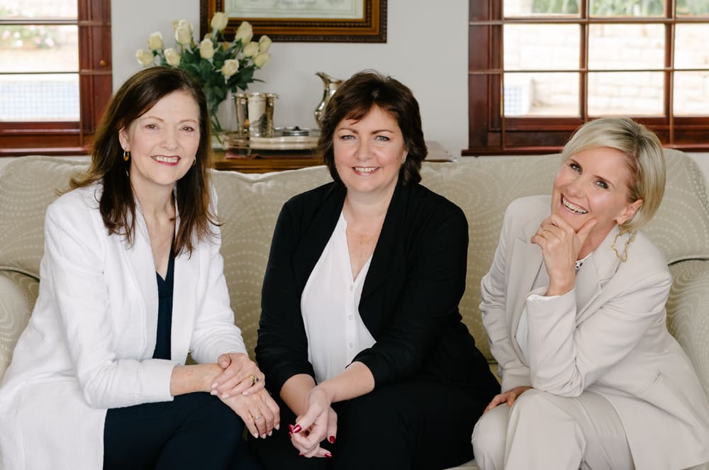 The image shows three women sitting together on a couch in what appears to be a cozy living room setting. They are all smiling and dressed in formal attire, suggesting a social or professional gathering.