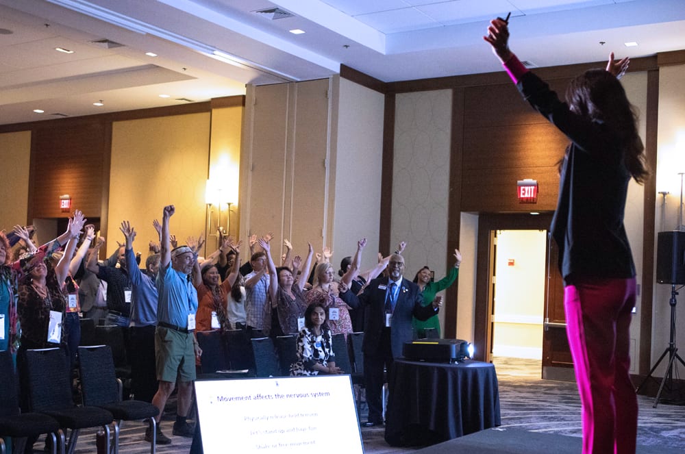 A large group of people with their arms raised, standing in a conference room or event space with tables and chairs in the background.