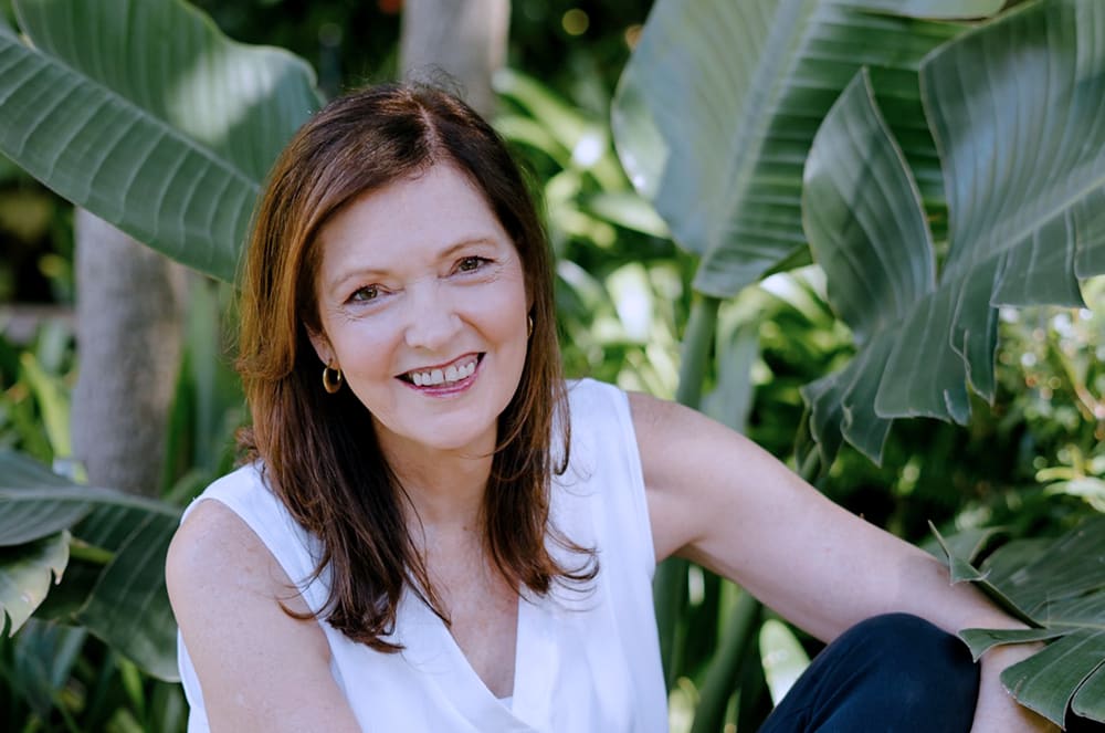Aletta Rochat, smiling woman with long brown hair sits in a lush, green outdoor setting surrounded by large tropical leaves.
