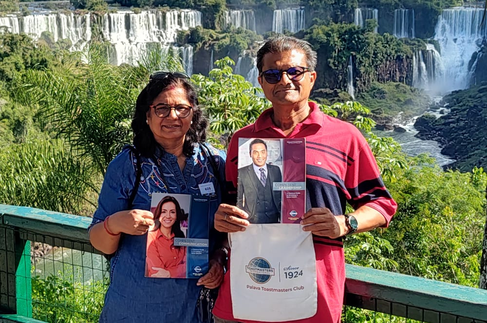 The image shows a couple standing in front of a scenic waterfall, holding framed portraits of what appears to be a political or public figure. A man and a woman holding Toastmasters products and standing outside in front of waterfalls