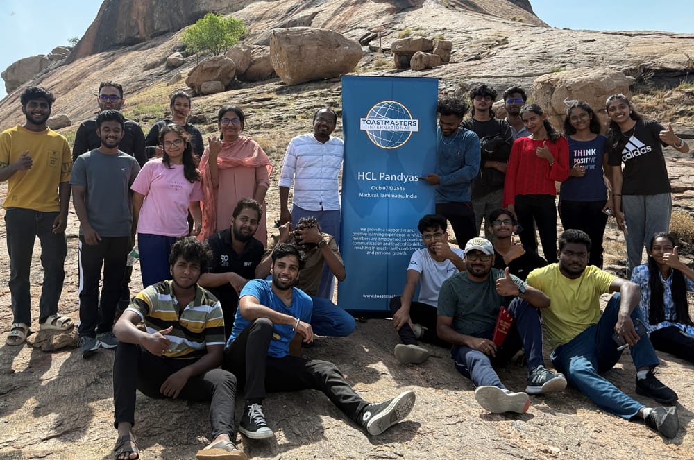 A group of people, mostly young adults, posing together in front of a large rock formation with a sign for HCL Pundits, an organization, in the foreground. Group of people posing around a blue Toastmasters banner outside