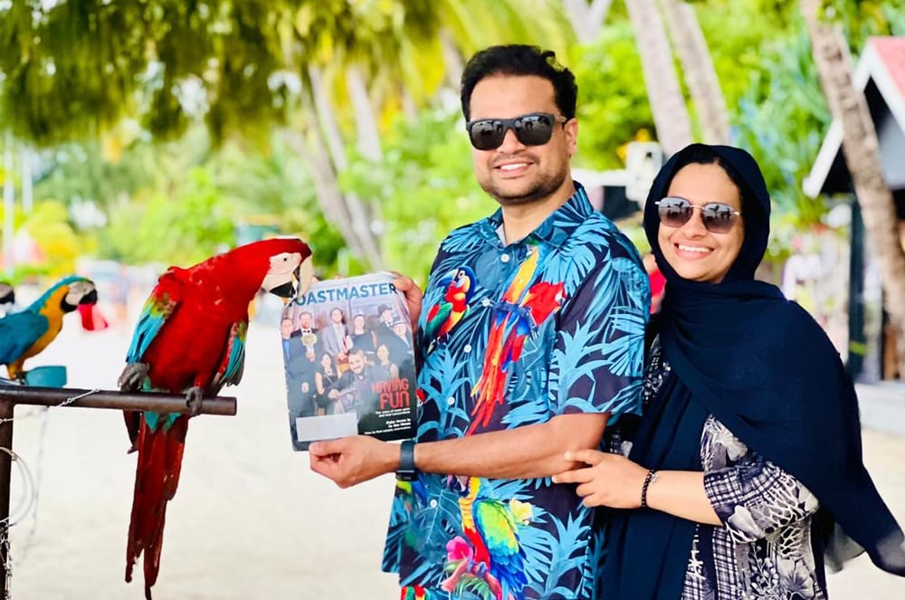 A smiling couple in tropical attire stands in front of a lush, green background with a colorful parrot perched nearby.