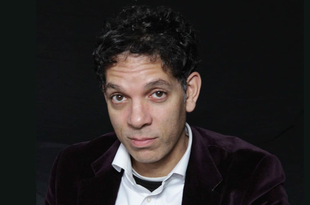 A man with dark, curly hair and an intense expression on his face is the focal point of the image, set against a dark background.