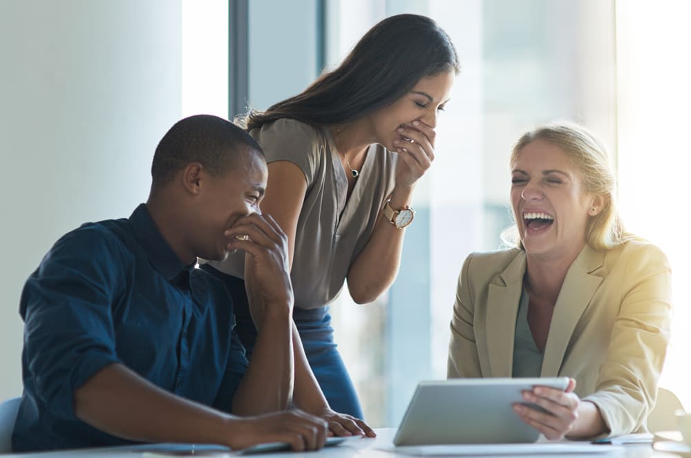 A group of three people, two women and one man, are gathered around a laptop, engaged in a lively discussion and sharing a moment of laughter.