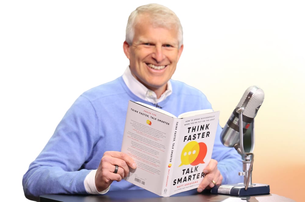 A smiling older man in a blue sweater is holding a book titled "Think Faster, Talk Smarter" while sitting in front of a microphone.