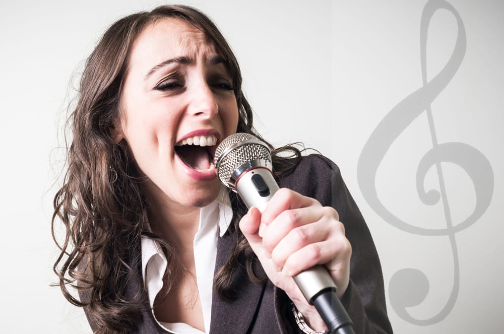 A young woman with long, dark hair is passionately singing into a microphone, with a musical note symbol visible in the background.