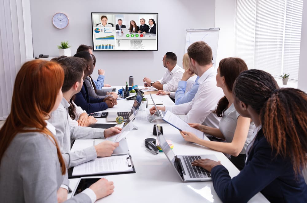 A group of people, likely professionals, are gathered around a table in an office setting, engaged in a meeting or discussion, with a large screen displaying data or information in the background.