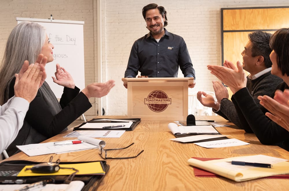 A group of people, including a man standing at a podium, are gathered in a meeting room, with various documents and office supplies visible on the table in the foreground.