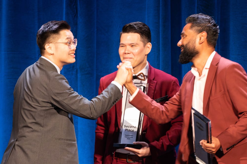 Three men in formal attire standing on a stage with a blue curtain backdrop, engaged in what appears to be a discussion or presentation.