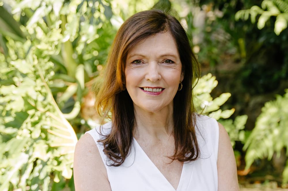 A smiling woman with long brown hair stands in a lush, green outdoor setting with trees and foliage in the background.