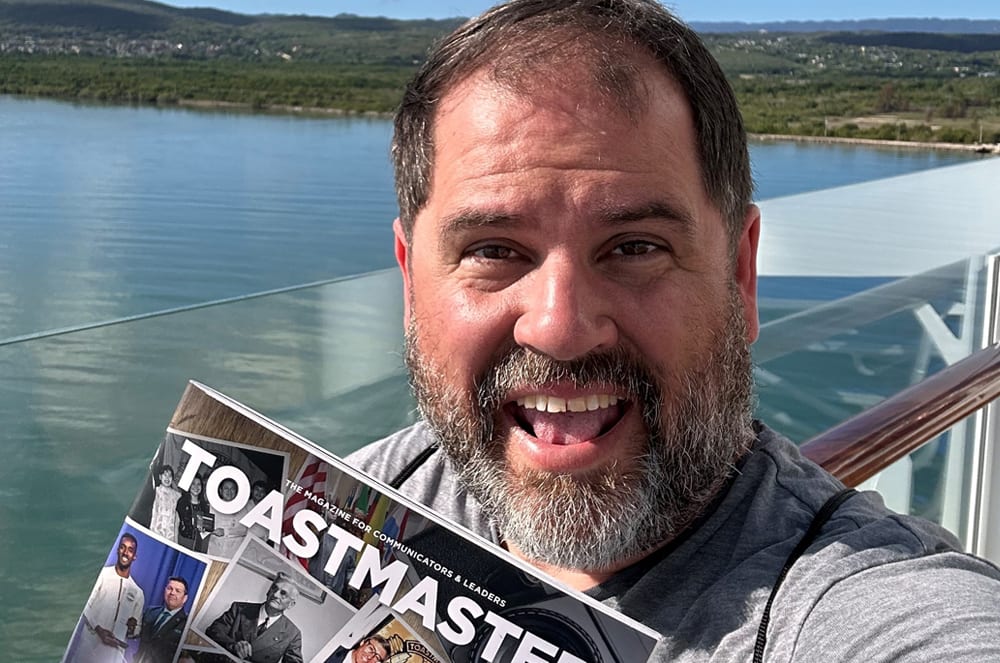 A bearded man with a joyful expression is holding a newspaper titled "Toastmaster" in front of a scenic lake and mountainous landscape in the background.