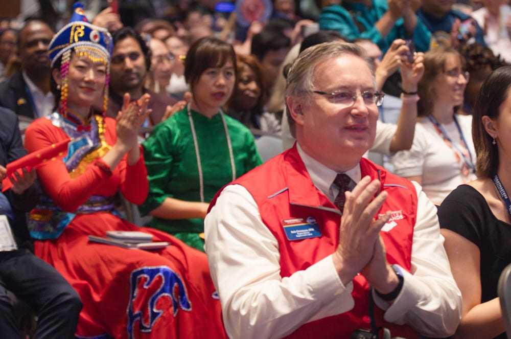A large crowd of people, some wearing colorful traditional clothing, are gathered together, with a man in a red vest standing in the foreground applauding.