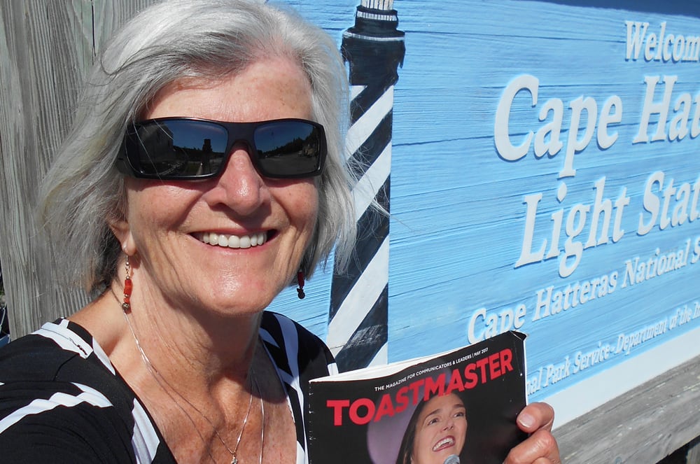 A smiling older woman wearing sunglasses and holding a magazine stands in front of a mural depicting the Cape Hatteras Lighthouse.