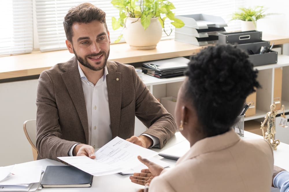 A well-dressed man is sitting at a desk, engaged in a conversation with a woman across from him, in an office setting with various office equipment and decor visible in the background.