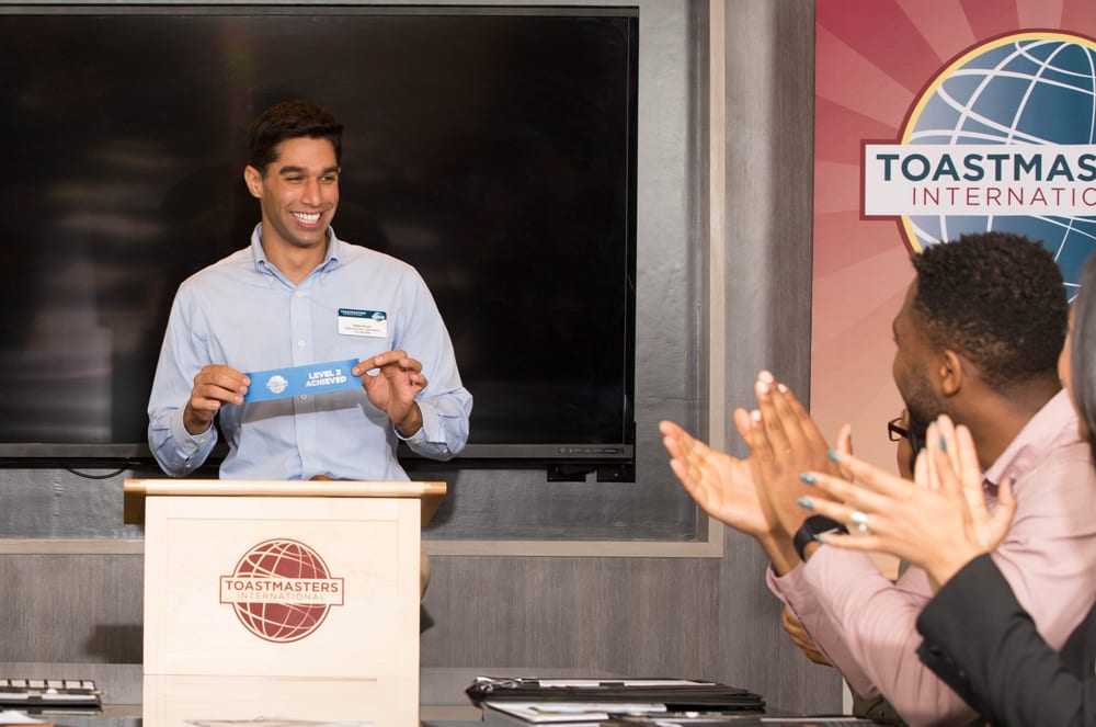 A man in a blue shirt is standing at a podium with a Toastmasters International logo, while another person is applauding him in the background.