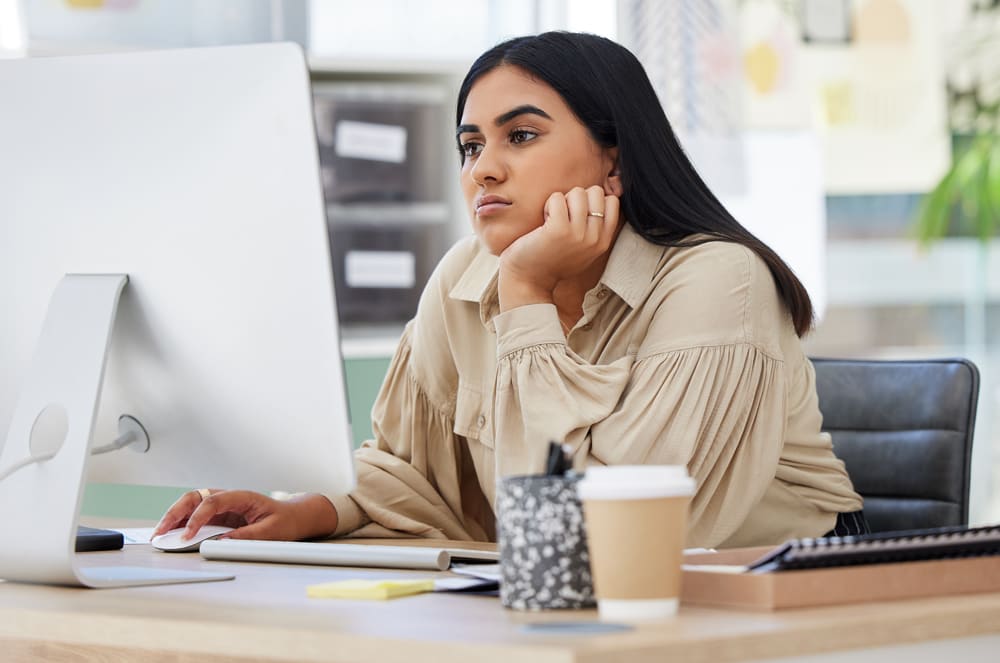 A young woman with long dark hair sits at a desk, working intently on a computer while holding a coffee cup.