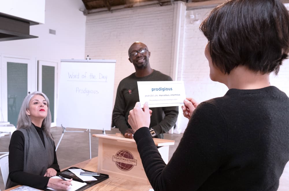 The image shows a group of people in a professional setting, with one person holding a sign that appears to be related to a presentation or event.