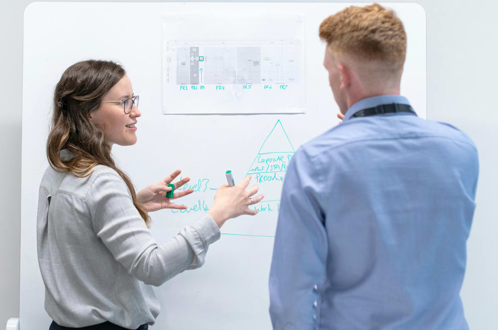Two people, a woman and a man, are standing in front of a whiteboard discussing something, with the woman gesturing and the man listening intently.