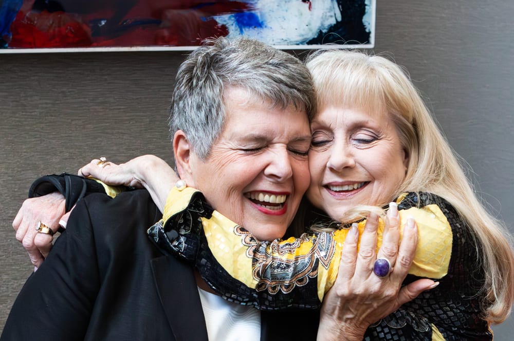 Two older women with gray and blonde hair are embracing and smiling joyfully in the foreground, with an abstract painting visible in the background.