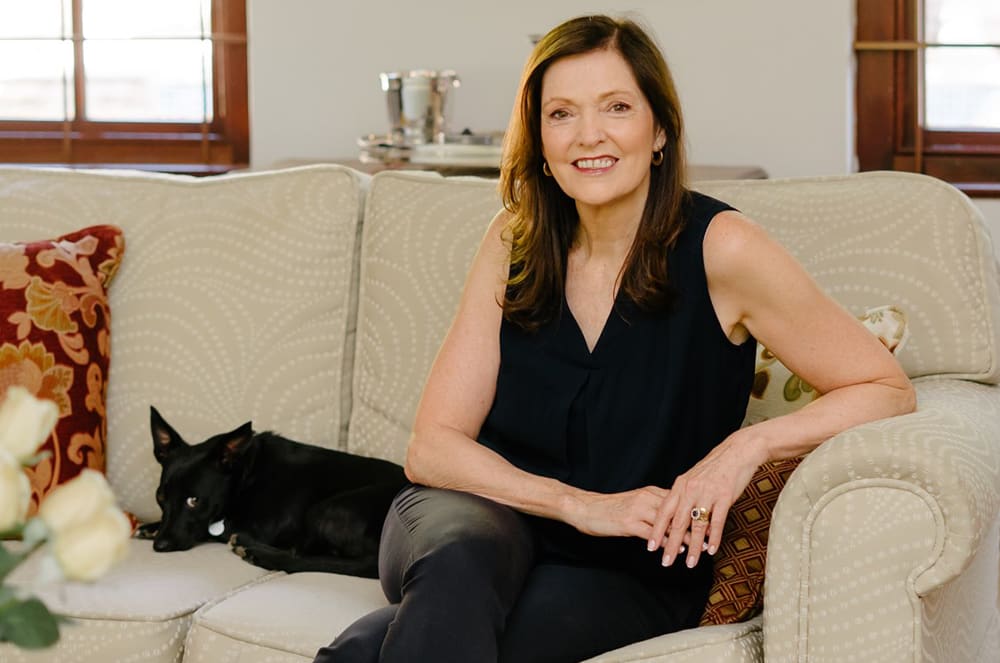 A woman with long brown hair is sitting on a couch, smiling and petting a black dog that is lying next to her. The room behind them appears to be a cozy living space with a window and decorative elements.