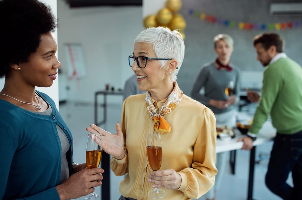 Two women in casual attire are engaged in conversation, with a group of people visible in the background.