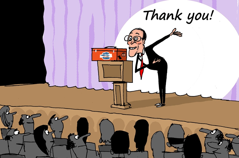 A man in a suit stands on a stage, waving his arms and thanking the audience seated in front of him.