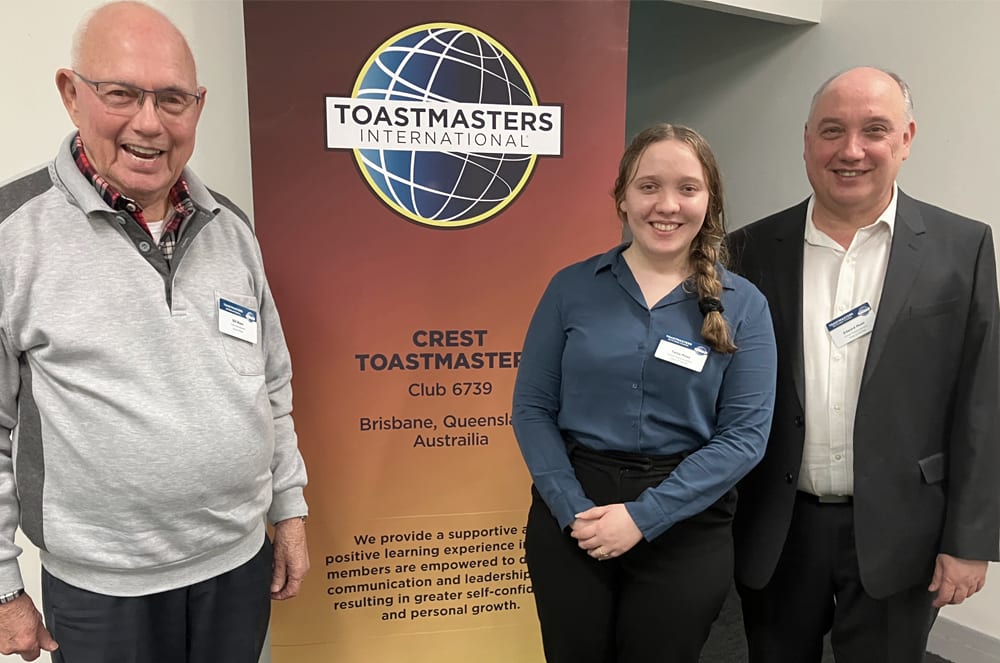 The image shows three people standing in front of a Toastmasters International banner, with the Crest Toastmasters club 6739 from Brisbane, Queensland, Australia displayed.