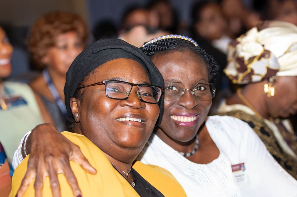 Two smiling women with glasses are embracing each other in the foreground, surrounded by a crowd of people in the background.